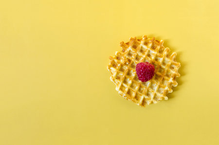 Waffle with a large raspberry berry on a yellow background close-up. Homemade crispy waffle on a colored backgroundの写真素材
