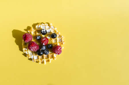 A waffle with different berries of black and red color on a yellow background is hard light. Homemade cakes crispy waffle on a colored backgroundの写真素材