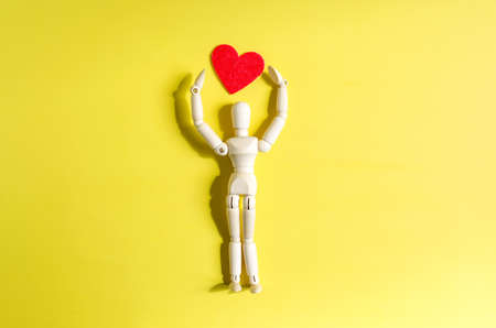 The figure of a wooden man with his hands raised above his head holding a heart on a bright background. Valentine's Day card man and heart on a yellow background close-up.の写真素材