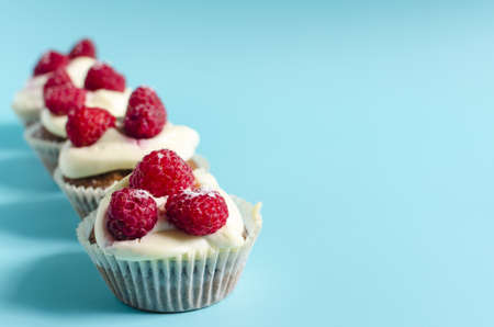 A row of cupcakes with white cream and raspberries on a blue background sharpness in the foreground. Sweet homemade cupcakes on a colored background are a place for text.の写真素材