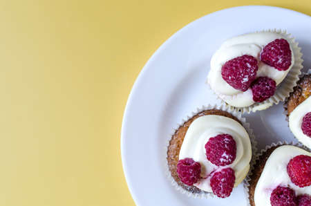 Cupcakes with white cream and raspberries in a white plate on a yellow background. Sweet homemade cakes close-up on a colored background.の写真素材