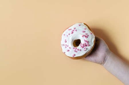 A hand holding a fresh donut with colored glaze on a beige background. The concept of white on a colored background is a place for close-up textの写真素材