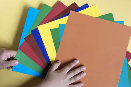 Children's hands hold sheets of colored paper on a colored background. The concept of children's creativity is handmadeの写真素材