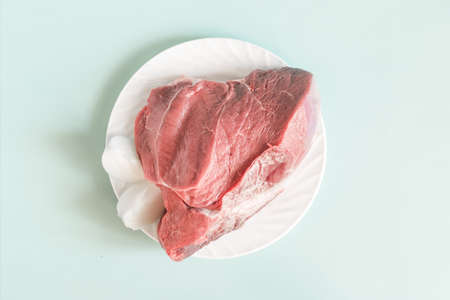 A piece of raw meat with onion slices on a white plate light blue background close-upの写真素材