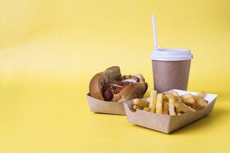 A fresh hot dog with sausage, French fries and a paper cup with a straw on a yellow copy space...