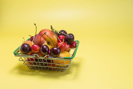 A large basket filled to the top with various fruits bananas, cherries, nectarines, apricots, peaches on a bright yellow background.の写真素材