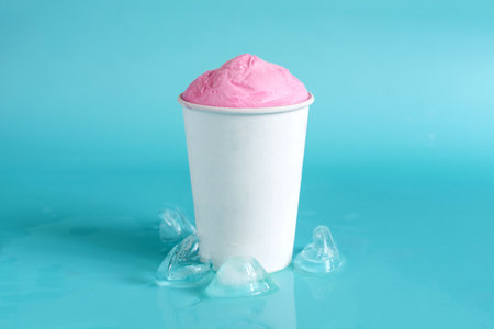 Pink ice cream in a white paper cup and ice cubes in close-up on a blue background.の写真素材