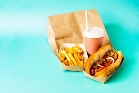 A paper bag, a fresh hot dog with sausage, French fries and a paper cup on a blue background close-up.の写真素材