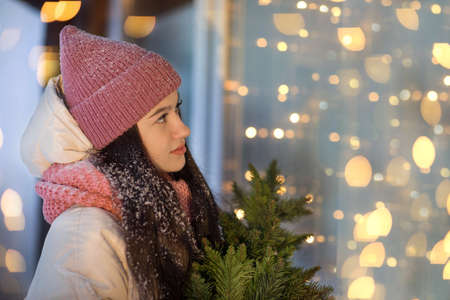 A beautiful girl stands near a decorated Christmas window, holding a Christmas tree in her hands, dressed in a hat and a warm scarf. High quality photoの写真素材