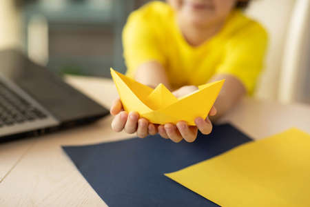 a boy in a yellow T-shirt at an online origami workshop, a paper boat. High quality photo