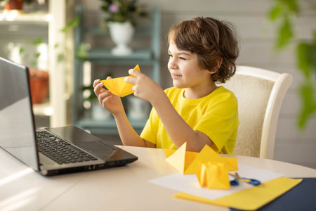 a boy in a yellow T-shirt at an online origami workshop, a paper boat. High quality photo