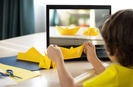 a boy in a yellow T-shirt at an online origami workshop, a paper boat. High quality photo