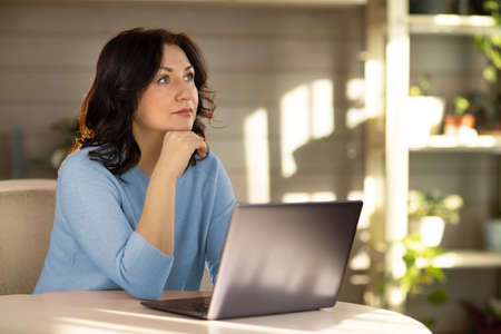 A 35-year-old woman in a blue jacket, sitting at the computer and looking wistfully to the side.の写真素材