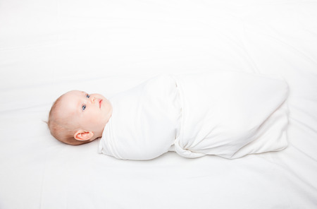 Three month baby girl swaddled in white blanket laying on a bed. Swaddling is a practice of wrapping infants in cloths in order to prevent limb movement. Medical and psychological effects of swaddling are controversial.の写真素材