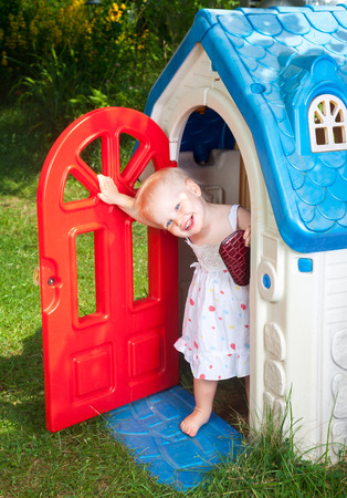 Little baby girl wearing white dress looking out from plastic play house doorway in a summer playgroundの写真素材