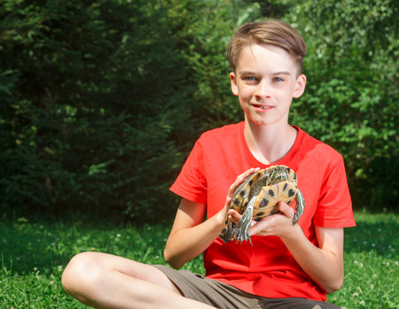 Cute teenager boy wearing red t-shirt sitting on a lawn in a summer garden holding turtle looking at camera smiling focus on turtleの写真素材