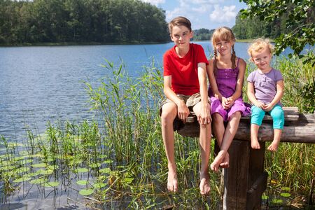 Three children of different age - teenager boy, elementary age girl and toddler girl sitting on a wooden pier by a forest lake looking at camera smilingの写真素材