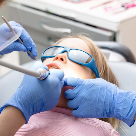Dentist performing dental filling procedure to a little girl in pediatric dental clinic. Doctor removing caries using high-speed dental drill. Calm child is sitting in a dental chair wearing sunglasses holding mouth openの写真素材
