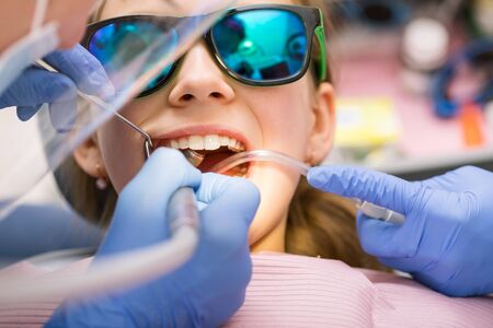 Dentist performing dental filling procedure to preteen girl in pediatric dental clinic. Doctor removing caries using high-speed dental drill. Child is sitting in a dental chair wearing sunglassesの写真素材
