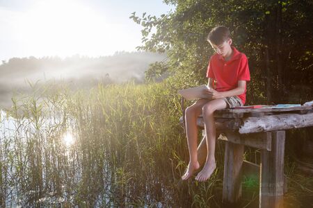 Teenager boy sitting on a wooden pier by forest lake drawing with pastel crayonsの写真素材