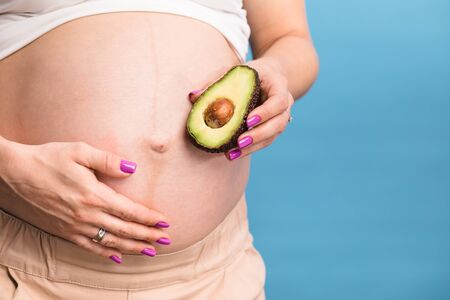 Studio shot of young pregnant woman holding one half of a fresh avocado fruit close to her belly...