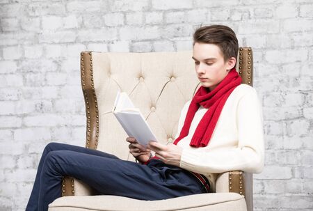 Teenager boy wearing red scarf and white sweater sitting in an armchair at home reading a bookの写真素材