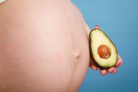 Studio shot of young pregnant woman holding one half of a fresh avocado fruit close to her belly...