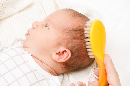 Mother brushing hair of her newborn baby using soft hairbrush to stimulate infant’s hair follicles...