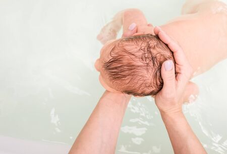 Mother bathing newborn baby supporting child's head and neck with her arms while baby is floating in bathtubの写真素材
