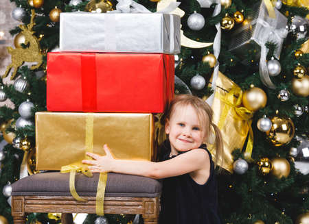 Portrait of elementary age girl wearing blue dress standing beside christmas tree holding pile of...