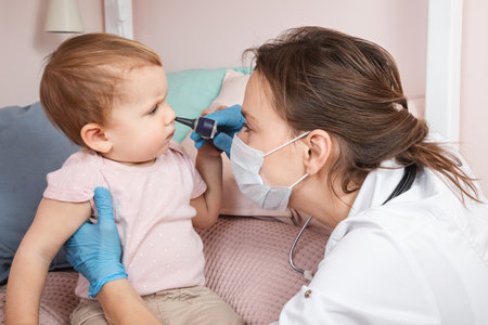 Pediatrician examines nose of baby girl at home during coronavirus COVID-19 pandemic quarantine. Doctor using otoscope (auriscope) to check nasal passage of a childの写真素材