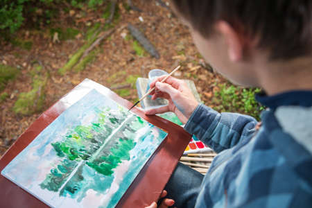 Teenager boy sitting by forest lake painting landscape with watercolor - plein air painting conceptの写真素材