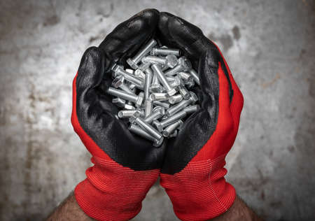 Person wearing protective gloves holding a handful of bolts and nuts in cupped hands against metal backgroundの写真素材
