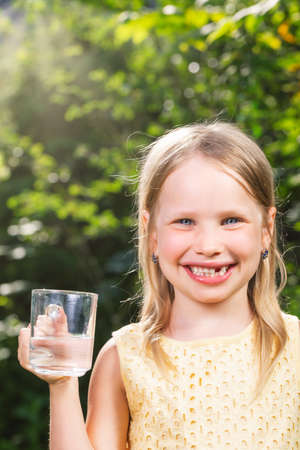 Happy little girl wearing yellow dress holding glass cup of water in a summer garden - healthy lifestyle conceptの写真素材