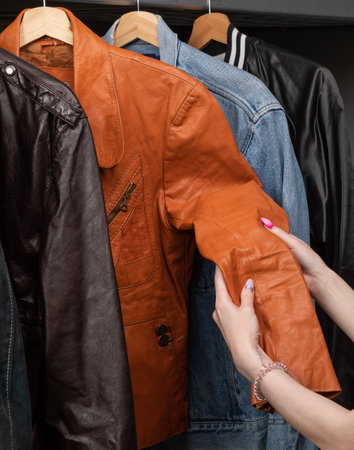 Woman choosing clothing in a second hand store. Various vintage suede leather and jeans jackets hang on clothing rack. Thrifting and sustainability in clothing conceptの写真素材
