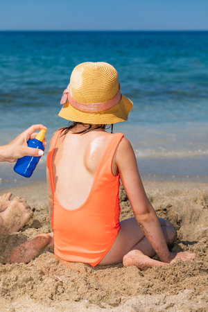 Mother spraying sunscreen on her daughter's back while she playing on a beach