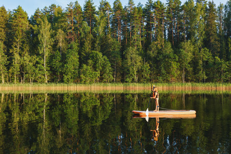 Little girl wearing life jacket paddling on a stand-up paddleboard (SUP) on a forest lake during the...
