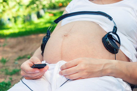 Young pregnant woman sitting in a lounge chair in a summer garden with music player and headphones on her bellyの写真素材