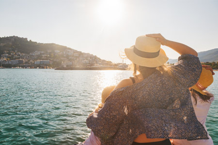 A mother and two children wearing straw hats seen from behind gazing at a scenic coastal city from a boatの写真素材