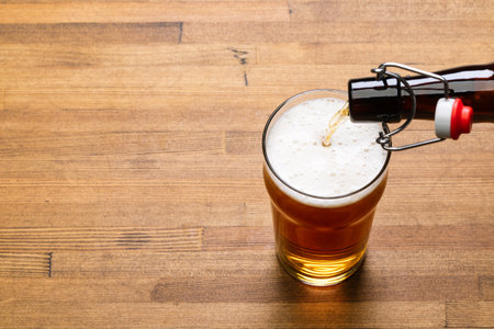 Craft beer is pouring from swing top bottle into nonic pint glass placed on a warm-toned wooden tableの写真素材