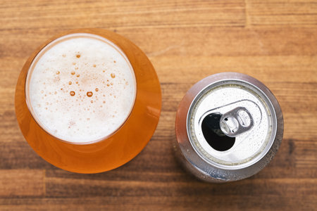 Top-down view of glass filled with a golden ale with an open beer can with condensation against rustic wooden tableの写真素材