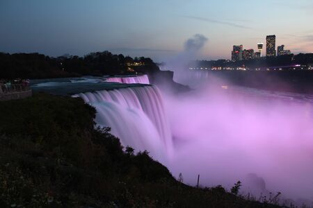 Wide view of Niagara Falls illuminated pink in evening as seen from USA, with water flow all blurred, cityscape visible on Canadaの写真素材