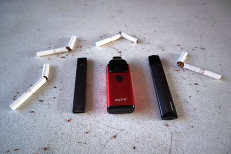 Different vape devices electronic cigarettes juul, aspire breeze, smok infinix, as smoking alternatives with broken marlboro gold cigarettes and scattered tobacco on white textured backgroundのeditorial素材