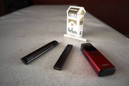 3 vapes juul, aspire breeze, smok infinix with a pack of marlboro cigarettes and one cigarette placed outside on a white textured table, isolatedのeditorial素材