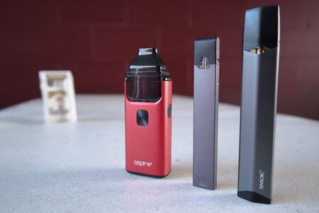 3 different vape pen electronic cigarette devices juul, aspire breeze, smok infinix, with a pack of Marlboro Gold cigarettes in the background out of focusのeditorial素材