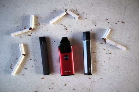 Different vape devices electronic cigarettes juul, aspire breeze, smok infinix, as smoking alternatives with broken marlboro gold cigarettes and scattered tobacco on white textured backgroundのeditorial素材