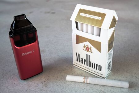 aspire breeze vape pod electronic cigarette device with a pack of marlboro cigarettes and one cigarette placed outside on a white textured table, isolatedのeditorial素材