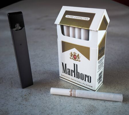 juul vape pod electronic cigarette device with a pack of marlboro cigarettes and one cigarette placed outside on a white textured table, isolatedのeditorial素材