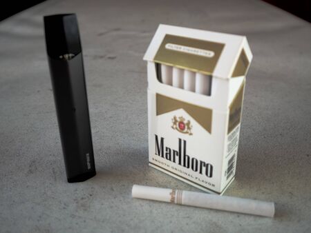 smok infinix vape pod electronic cigarette device with a pack of marlboro cigarettes and one cigarette placed outside on a white textured table, isolatedのeditorial素材