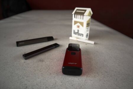 3 vapes juul, aspire breeze, smok infinix with a pack of marlboro cigarettes and one cigarette placed outside on a white textured table, isolatedのeditorial素材